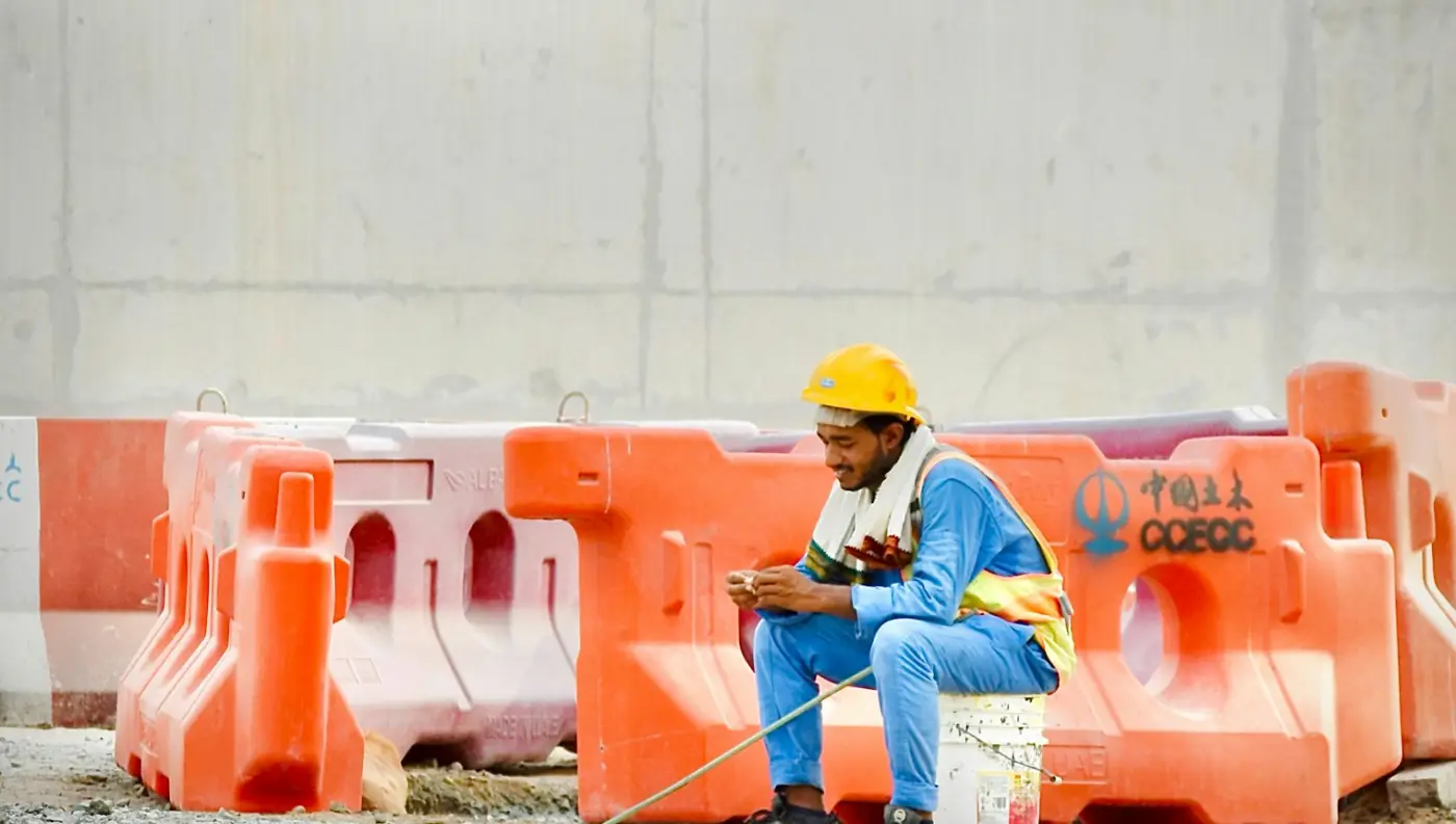 Construction worker in a hard hat sitting on an upturned bucket, checking his phone on a break